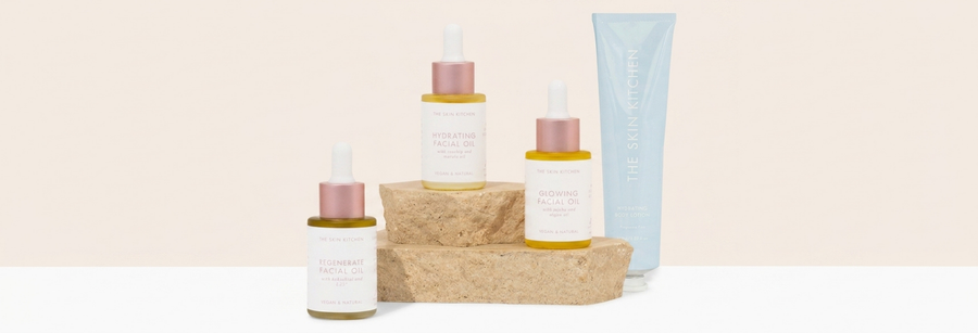 Product lineup of The Skin Kitchen skincare items including facial oils and body lotion arranged on stone pedestals against a light beige background.
